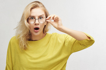 Surprised woman in round glasses with open mouth and bulging eyes looks into camera and sees something incredible and amazing isolated on white background - obrazy, fototapety, plakaty