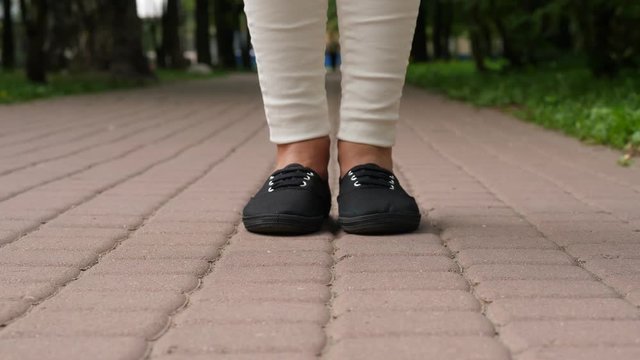 Young adult woman in black light sneakers and white jeans stay at pavement. Low camera position, only bottom of legs and shoes shown on tile paving. Girl stay still with feet close. Blurred background