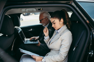 Good looking senior business man and his young woman colleague or coworker sitting on backseat in luxury car. They talking, smiling and using laptop and smart phones. 