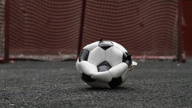 Broken half deflated soccer ball lie on black rubber field against small gate seen blurred on background. Worn out ball looks relatively clean and new, but crumpled and shabby