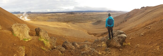 Young man wearing a blue jacket stands on top of a mud mountain, overlooking a geothermal spot noted for its bubbling pools of mud & steaming fumaroles emitting sulfuric gas. Power of planet Earth.