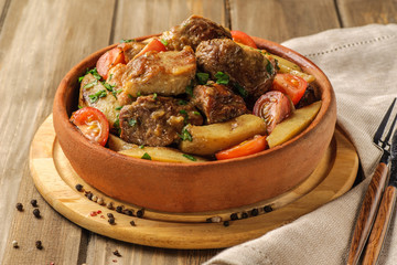 The traditional pork and potato dish of Georgian cuisine on earthenware ketsi. Ojahuri with tomatoes, hot peppers and fresh herbs. Dark background and natural napkin. Horizontal view.