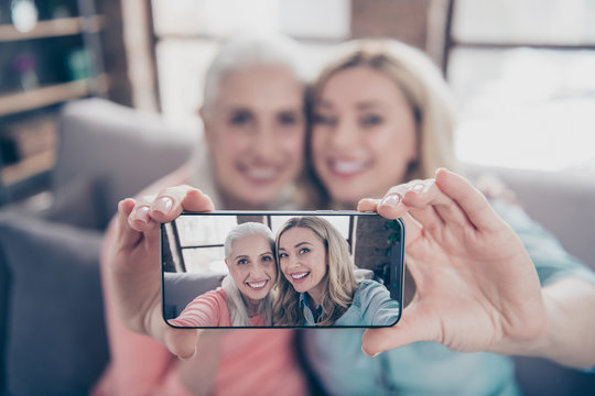 Close up photo two beautiful she her ladies funky granny granddaughter relatives mom mommy make take selfies toothy telephone hands arms blurred picture sit couch divan sofa house indoors