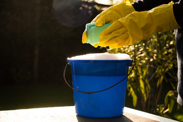 Spring Cleaning outside with big yellow cleaning gloves, water, soap and a big blue bucket with soap. Cleaning the table. Soap Foam on the table. Hands in bucket. Holding and wringing cleaning sponge. © Pepijn