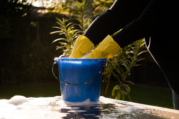 Spring Cleaning outside with big yellow cleaning gloves, water, soap and a big blue bucket with soap. Cleaning the table. Soap Foam on the table. Hands in bucket. Holding and wringing cleaning sponge. © Pepijn