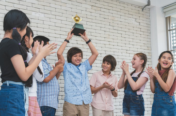 Group of children congratulated the happy boy holding trophy cup award of his winning in classroom
