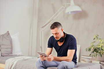 Caucasian man in forties sitting on bed in bedroom and using tablet.