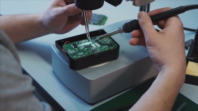 Hands of professional tech repairing air drone components on microscope table using soldering iron. Closeup. On the printed circuit board are visible traces of short circuit, burnt