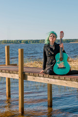 A musician with blue hair and a blue guitar sits on a lake footbridge and plays music.