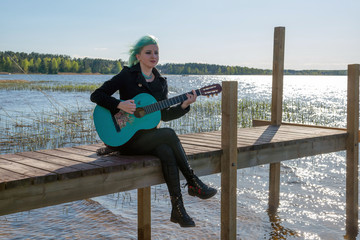A musician with blue hair and a blue guitar sits on a lake footbridge and plays music.
