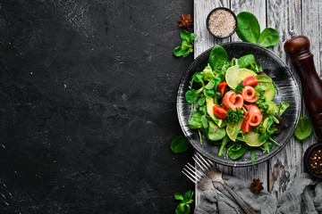 Salad with salmon, avocados, tomatoes and spinach. In a black plate on a wooden background Top view. Free space for your text. Flat lay