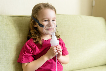 Little girl holds inhaler mask at home. Sick kid breathes through a nebulizer. Baby using equipment...