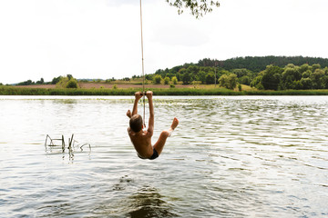 Teenage boy jumping in the river from the swinging rope on sunny summer day.