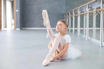 Little ballerina is trying on pointe shoes in ballet class. Cute child girl is sitting on floor...