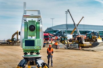 Surveyor engineer with equipment (theodolite or total positioning station) on the construction site of the road or building with construction machinery background
