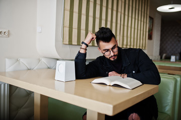 Arab man wear on black jeans jacket and eyeglasses sitting in cafe, read book. Stylish and fashionable arabian model guy.