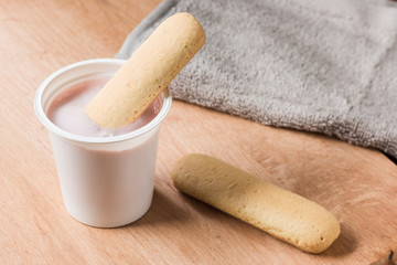 Fruit yogurt in a white plastic cup with biscuit cookies on a wooden kitchen table.