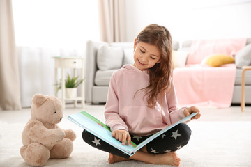 Cute little girl with teddy bear reading book on floor at home