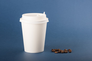White disposable paper cup with white cap and coffee beans on a blue background