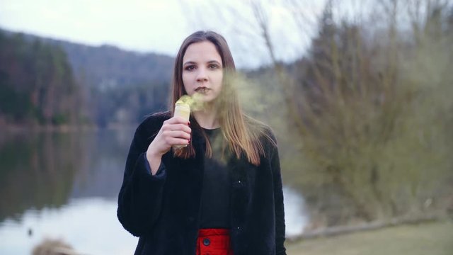 Young woman with a smoke bomb 4K. Static slow-motion shot of a female person in focus looking into the camera and holding a smoke bomb in one hand. Background lake and trees.