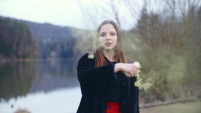 Girl playing with yellow smoke bomb 4K. Static slow-motion shot of a female person in focus spinning around and holding a smoke bomb in one hand. Background lake and trees.