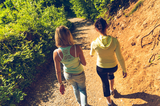 Two Girls Walking Together, View From Behind Following Young People Walking Through The Forest On Hiking Path