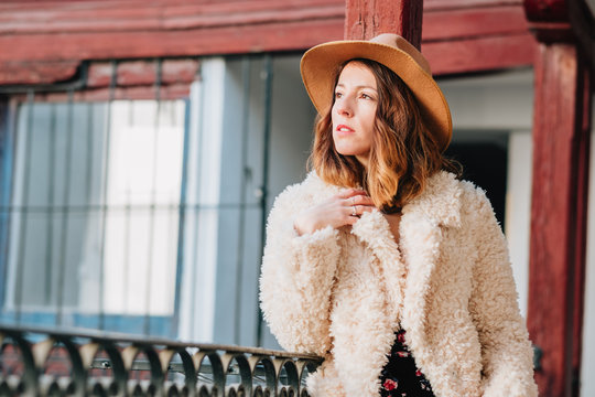 Positive attractive young lady in warm wear and hat looking away and standing near house and fence