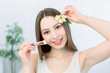 Beauty Woman face Portrait. Beautiful Spa model Girl with Perfect Fresh Clean Skin. Brunette female looking at camera holding makeup brush in one hand and the flower in another hand and smiling.