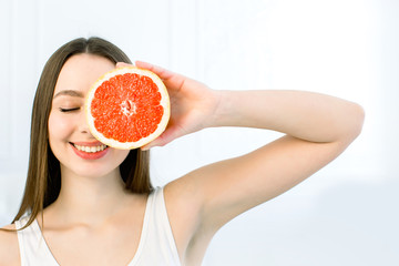 Portrait of young beautiful woman with healthy perfect skin holds piece of grapefruit, closing one eye. Natural cosmetics, skincare, wellness, facial treatment, cosmetology concept. Copy space