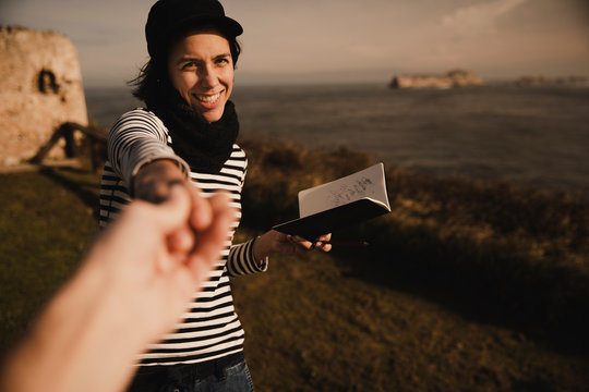 Elegant cheerful lady in cap with notepad holding crop hand of person on coast near sea