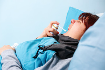 Image of sick brunette using throat spray lying in bed .