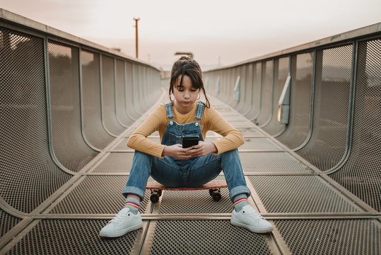 Stylish teen in jean overall sitting on skateboard and using mobile phone on footpath in city