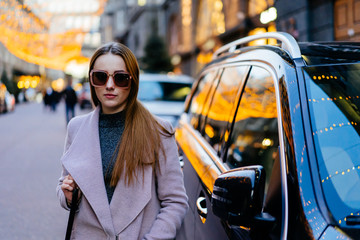 Fototapeta premium Beautiful young caucasian woman standing next to luxury SUV car in city downtown. Stylish female model wearing sunglasses photographed near expensive car.
