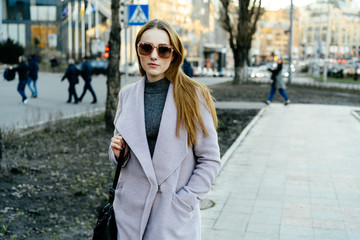 Beautiful young caucasian woman walking along the street in the city. Stylish female model wearing sunglasses walking on city street.