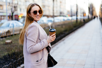 Beautiful young caucasian woman walking along the street in the city. Stylish female model wearing sunglasses with disposable cup walking on city street.