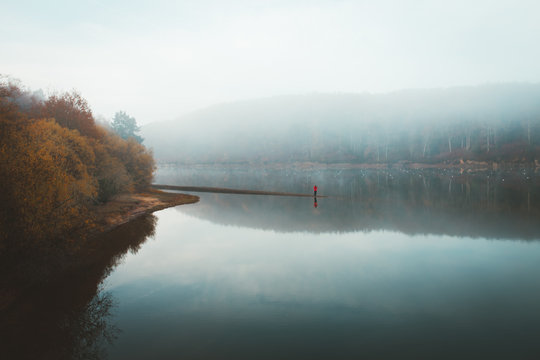 Picturesque landscape of beautiful silent lake with reflection of forest and lonely tourist in thick fog