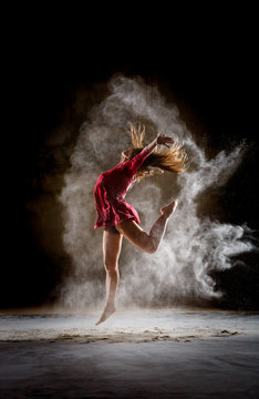 Side view of young slim ballerina in red dress upping leg and hands between mist in dark room