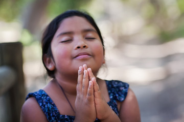 Fototapeta na wymiar A native young girl with hands together in prayer, in an outdoor setting praying to God with a subtle smile.