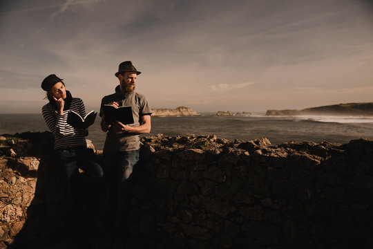 Stylish guy in hat and elegant lady in cap reading volumes near rocks on coast of sea and wonderful sky
