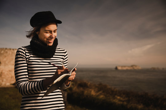 Elegant lady in cap taking notes in notepad on seat on coast near waving sea