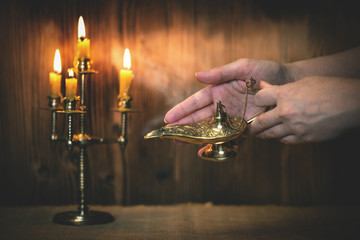 A golden Genie lamp in a woman hands on a wooden board background in the light of burning candle....