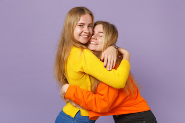 Two pretty joyful young blonde twins sisters girls in vivid colorful clothes hugging looking camera isolated on pastel violet blue wall background. People family lifestyle concept. Mock up copy space.