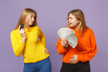 Two shocked blonde twins sisters girls holding credit bank card, fan of money in dollar banknotes, cash money isolated on violet blue background. People family lifestyle concept. Mock up copy space.
