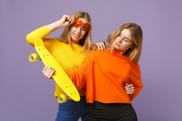 Two stunning young blonde twins sisters girls in vivid clothes, heart eyeglasses hold yellow skateboard isolated on pastel violet blue background. People family lifestyle concept. Mock up copy space.