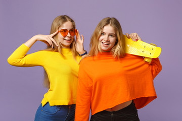 Two attractive young blonde twins sisters girls in vivid clothes heart eyeglasses hold yellow skateboard isolated on pastel violet blue background. People family lifestyle concept. Mock up copy space.