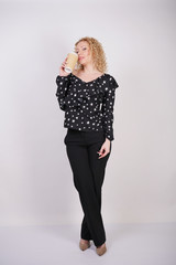cute curly blonde girl stands in black fashionable clothes and holds a paper Cup of coffee on a white Studio background