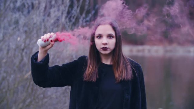 Rebellious woman in black with smoke walk towards the camera 4K. Static slow-motion shot of a female person in focus looking into the camera and holding a smoke bomb in one hand. 