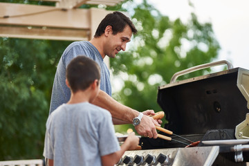 father and son grilling hot dogs together on backyard gas grill - obrazy, fototapety, plakaty
