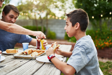 happy hispanic family eating grilled hot dogs on picnic table in backyard