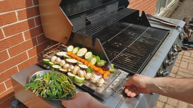 Home cooking - Placing on gas grill side table a stainless steel tray of skewers of assorted vegetables and dish of green beans and asparagus.